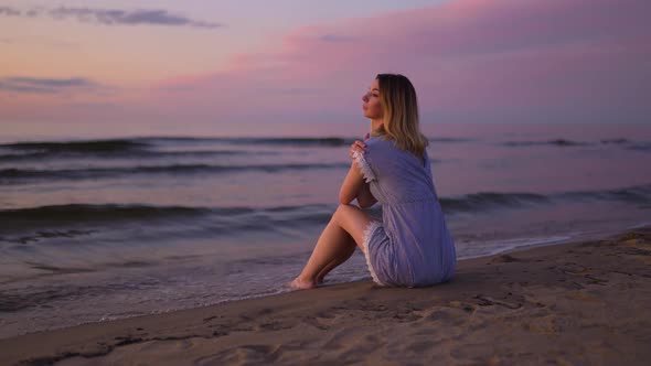 Backside View Young Woman with Naked Legs Sitting at Sea on the Beach Wearing Blue Summer Dress alt