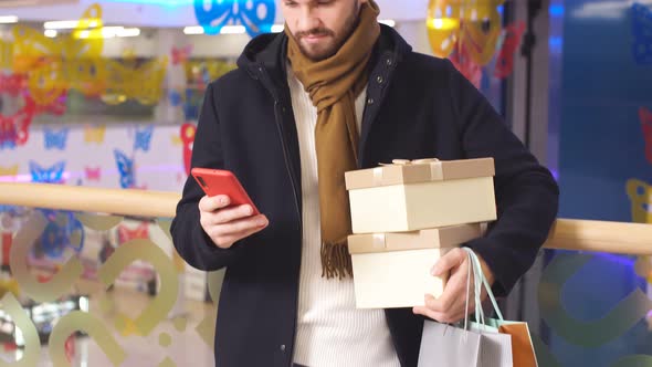 Young Cute Men in Dark Coat Uses Smartphone in the Interior of Shopping Center alt