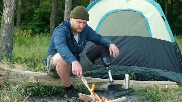 Disabled man with prosthetic leg is sitting on wooden log in forest near fire. alt