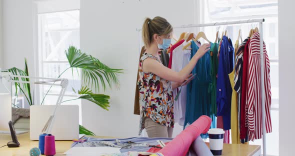 Female fashion designer wearing face mask looking checking on dresses at studio alt