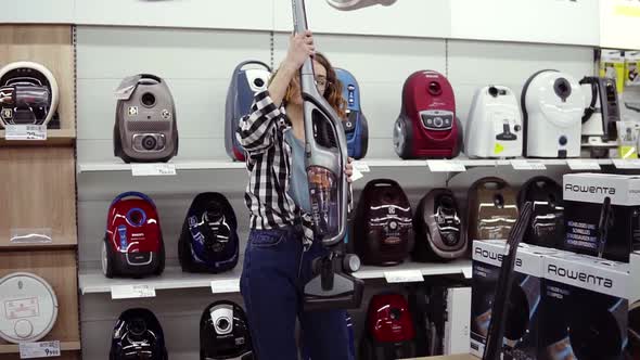 Cheerful Curly Haired Woman Standing Near New Vacuum Cleaners in Home Appliance Store Choosing the alt