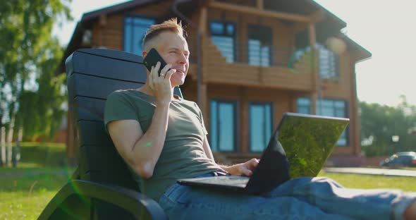 Happy Young Man Sitting on Background of Country House with Laptop and Talking on the Phone alt