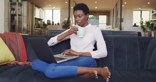 Happy african american woman sitting on sofa in living room, using laptop alt