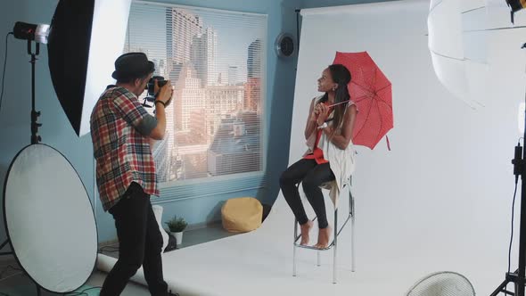 Stylish African Model Posing with Red Umbrella on Bar High Chair for Fashion Magazine Photo Shoot alt