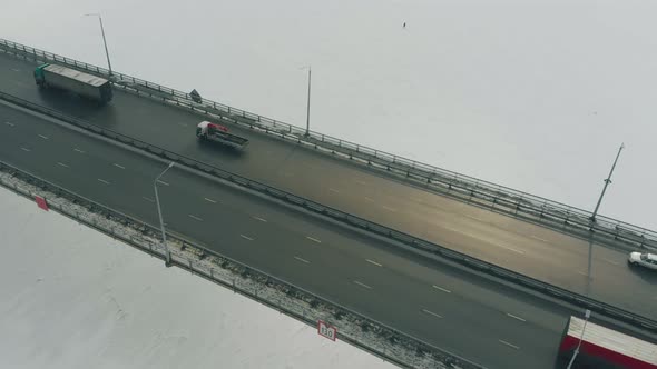 Trucks and Cars Drive Along Gray Bridge Over Frozen River alt