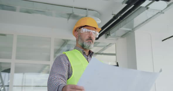 Builder in Special Uniform Holding Blueprint in Unfinished Office and Checking all Details alt