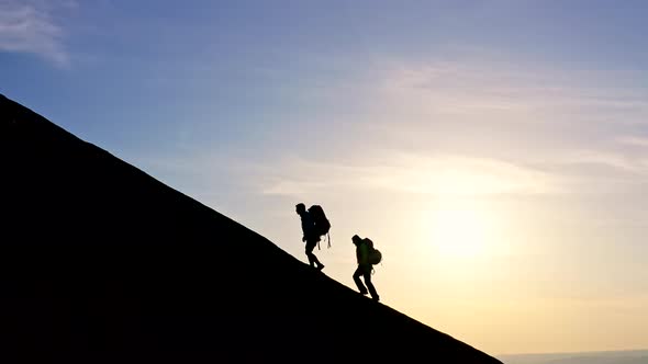 Silhouettes of a Man and a Woman with Backpacks Climbing to the Top of the Mountain