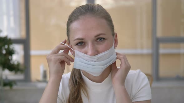 Cheerful Lady with Blue Eyes Puts on Disposable Mask in Cafe alt