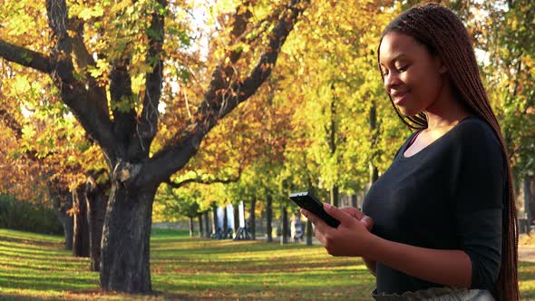 Young Beautiful Black Woman Works on Smartphone in Autumn Park - Falling Leaves alt