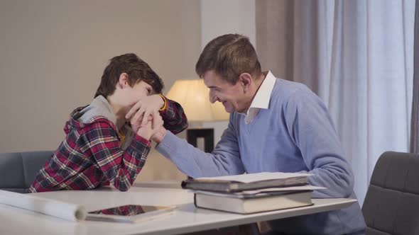 Cheerful Caucasian Boy Winning Middle Aged Grandfather in Arm-wrestling and Showing Victory Gesture alt