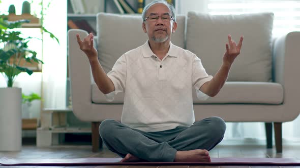 Asian elderly senior man doing yoga for meditation on yoga mat in living room alt