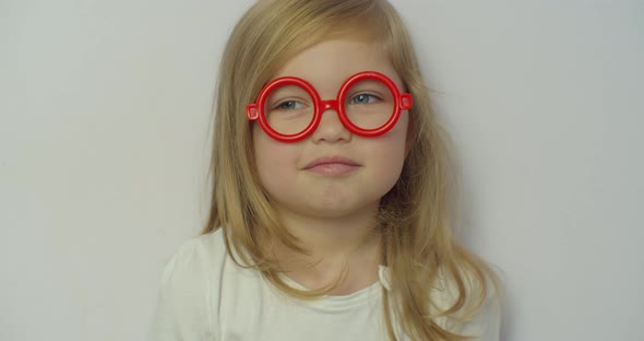 Close-up Little Blond Girl in Looking at Camera in Red Toy Glasses Stretches Smile on White alt