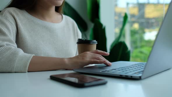 Closeup of a woman touching on laptop touchpad and drinking coffee with mobile phone on the table