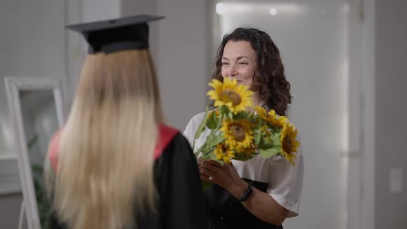 Smiling Happy Proud Mother Giving Bouquet of Yellow Sunflower to Graduate Daughter Talking alt