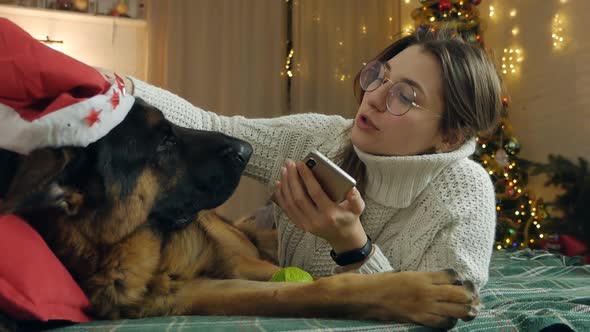 Young Woman Spend Christmas Holidays at Home with Her Pet on the Bed with Phone