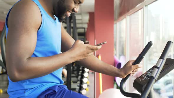 Joyful Male Athlete Riding Stationary Bike and Answering Phone Message in Gym alt