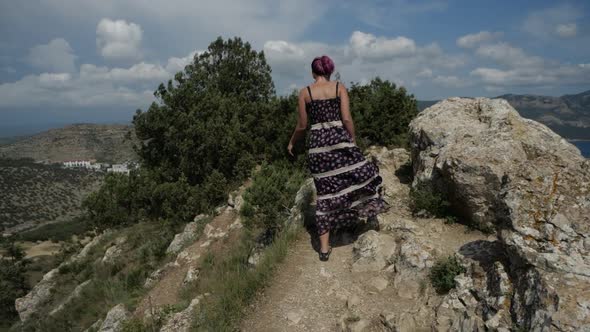 A Girl in a Long Dress Walks Up To the Top of a Mountain Overlooking the Sea Bay