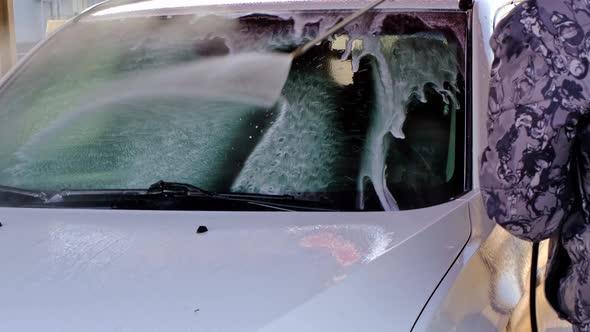 A man washes a white car at a self-service car wash with a high-pressure pistol. Washing off shampoo alt