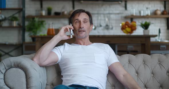 Smiling Senior Man Sitting Down on Couch at Living Room and Talking on the Smartphone alt