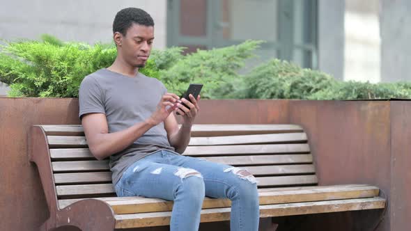 Young African Man Using Smartphone on Bench  alt