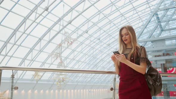 Happy Teenage Girl Holding Bags with Purchases, Smiling While Looking at Phone in Shopping Center alt