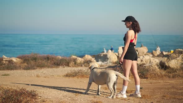 Wide Shot Confident Young Woman Pointing Away Standing with White Labrador on Mediterranean Sea alt