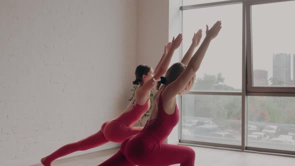 Two Women in Red Sports Uniforms Make Virabhadrasana in Yoga Studio alt