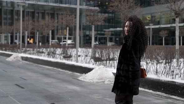 A Curlyhaired Woman in a Fur Coat Walks Through a Modern City Enjoying a Beautiful Day alt