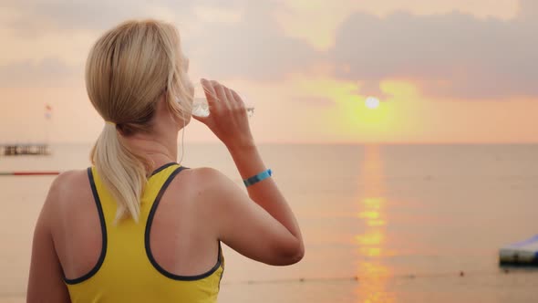 Active Woman Drinking Clean Water After Playing Sports. On the Background of the Sea, Where the Sun alt