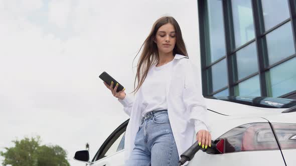 Young Woman is Standing Near the Electric Car and Looks at the Smart Phone alt