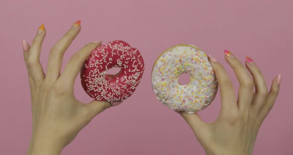 Womens Hands Holding Two White and Red, Sprinkled Donuts on Pink Background. alt