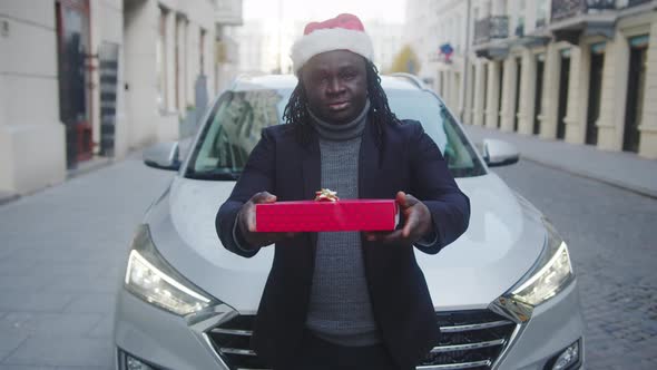 Young Black Man with Santa Hat Giving Red Wrapped Christmas Gift in Front of His Car alt