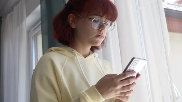 Young redhead ukrainian teen girl with mobile phone near window