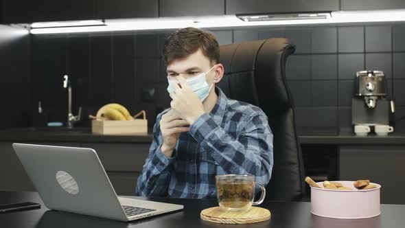 Young man puts on protective medical face mask and starts working on laptop at home. alt