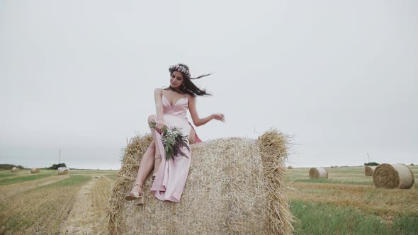 Happy Elegant Lady Sits on a Large Haystack in a Windy Field and Smiles alt