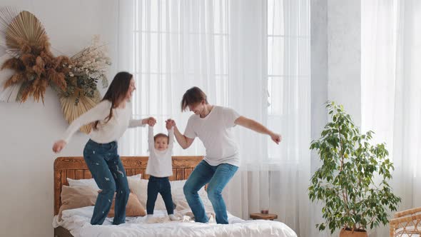 Young Parents Married Couple Caucasian Family with Little Daughter Toddler Child Girl Jumping on Bed alt