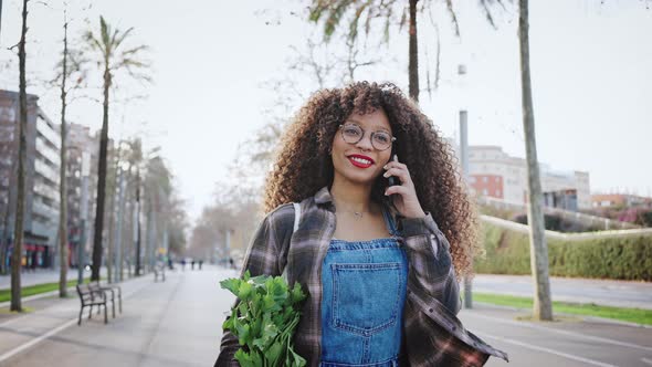 Mixed Race Lady with Long Curly Hair is Holding Mobile Phone While Walking Along Urban Street alt