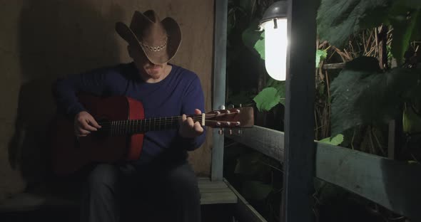 Man on Dark Night with Guitar is Sitting on Porch and Playing an Instrument