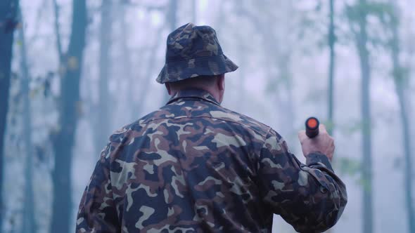 Back View of A Man in Camouflage Is Walking Through the Forest, Lighting His Way in the Fog alt