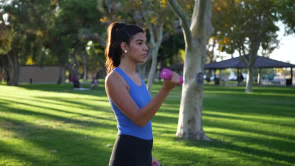 A young woman doing a dumbbell workout in the park performing bicep curls to build arm muscle and st alt