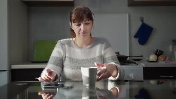 Happy Woman Sitting in the Kitchen at Home with a Cup of Tea and Uses the Phone, Play a Game