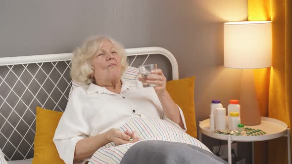 Sick Senior Woman Taking Medicines and Drinking Water While Lie on Bed Alone