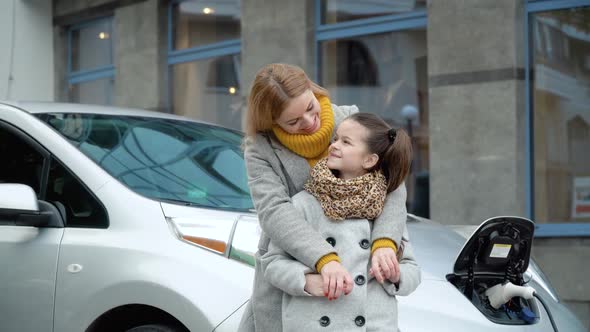 A Woman with Her Daughter Stands Near Her Electric Car and Looks at the Camera. Charging an Electric alt
