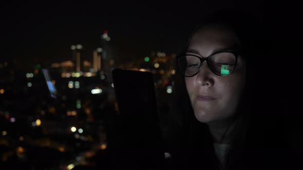 Sleepy Woman Is Flipping Pages on Phone at Night Near the Window with City View. alt
