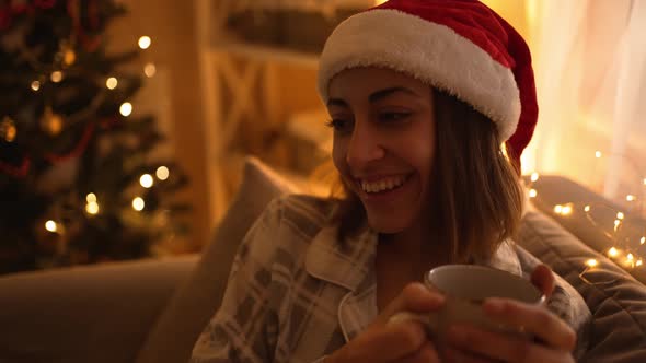 Woman in Pajama and Red Santa Hat Sitting on Couch Watching TV and Drinking Hot Chocolate From Mug alt