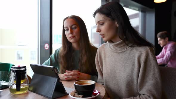 Two Young Beautiful Girls in a Cafe Discuss Work Issues Through a Tablet on a Zoom Conference