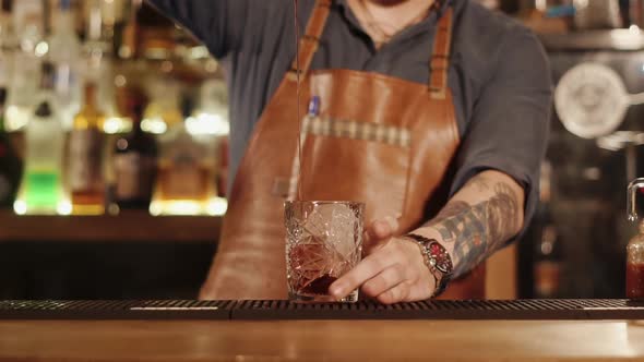Barman Is Pouring Mixed Drink in a Glass with Big Piece of Ice in a Bar alt
