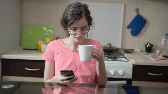 Girl with a Mobile Phone Sits in the Kitchen and Drinks Hot Tea alt