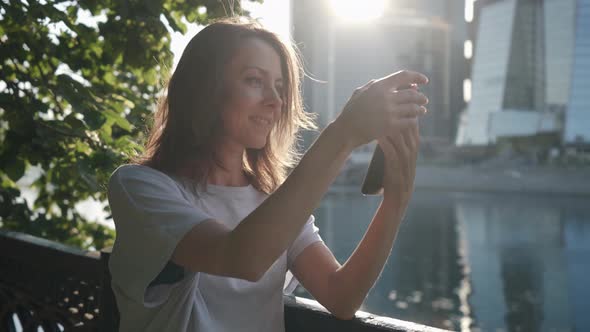 a Woman Stands with a Phone Pokes at the Screen and Smiles on the Embankment Against the Background alt