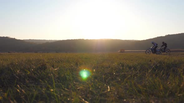 Two Motorcyclists Passing Through Large Field with Beautiful Sunset at Background alt